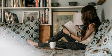 young woman seated on floor cushions with a hot drink in a mug, reading from a book, looking very cosy and relaxed