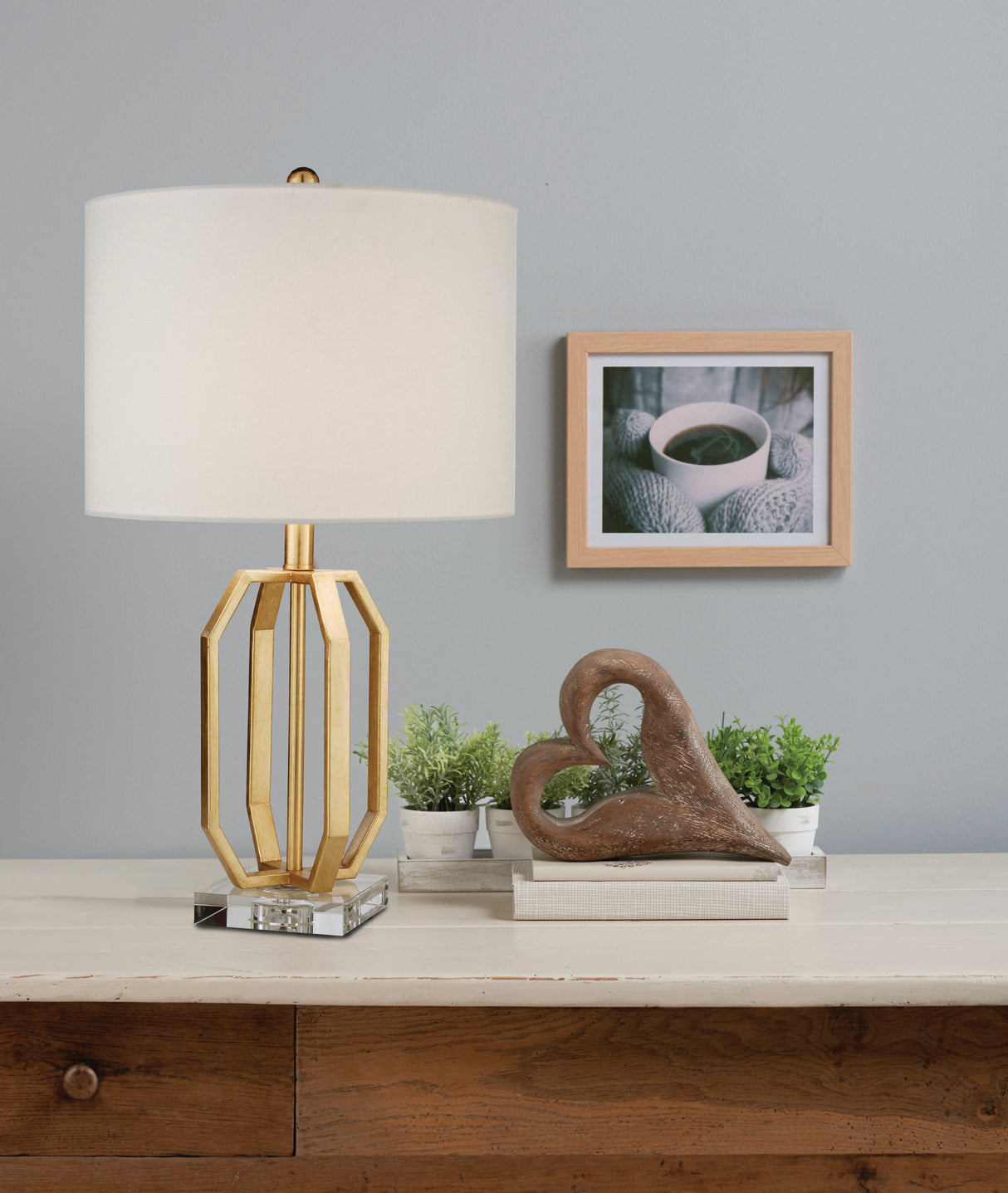 A cage style metal table lamp base on a square acrylic base plinth with a white straight sided cotton lampshade, stood on a wooden furniture unit in front of a pale blue wall. On the wall is a framed photo of a cup of coffee held in mittened hands. On the table to the right of the lamp is a brown wooden heart shaped ornament and potted plants.