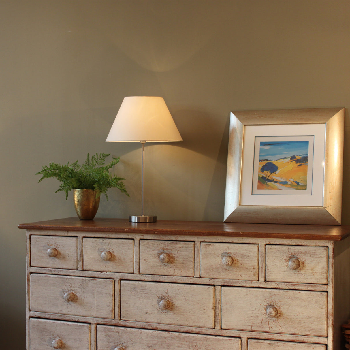 A picture of a wooden furniture unit in front of an olive green coloured wall. On top of the unit stands a green fern in a gold plant pot, a chrome metal table lamp base with a cream tapered lampshade and a gold framed picture. The lamp is turned on in this picture and you can see shadows and light on the wall behind.