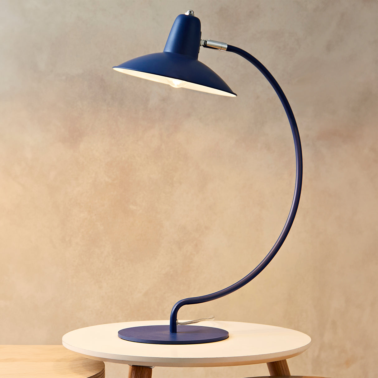 An arc shaped metal table lamp in a matte blue painted finish. The lamp is shown with a bulb just visible and is turned on in this picture. The lamp is placed on a small coffee table against a mottled beige coloured background.