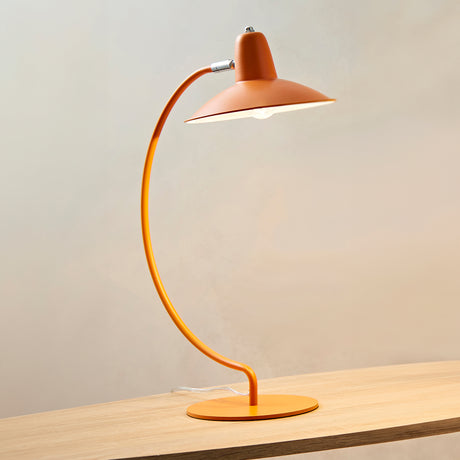 An arc shaped metal table lamp in a matte orange painted finish. The lamp is shown on a wooden table with a bulb just visible and is turned on in this picture.