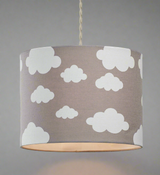 A picture of a grey straight sided cotton cyilnder lampshade with white printed cloud shapes in varying sizes. The picture is taken against a grey background and the bulb inside the pendant shade is turned on, casting a glow underneath the lampshade.