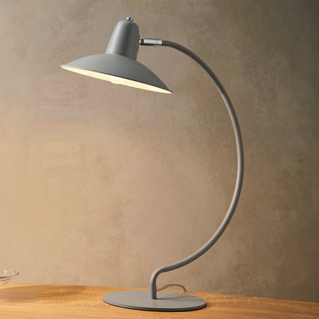 An arc shaped metal table lamp in a matte grey painted finish stood on a wooden table. The lamp is shown with a bulb just visible and is turned on in this picture.