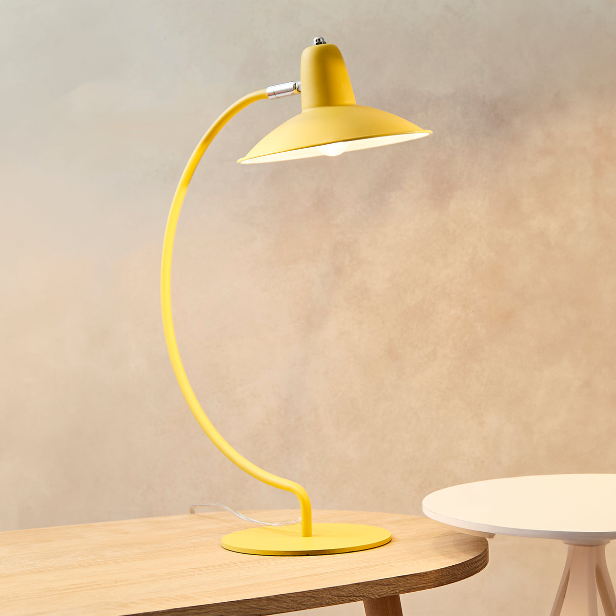 An arc shaped metal table lamp in a matte orange painted finish. The lamp is shown on a wooden table with a bulb just visible and is turned on in this picture.