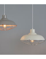 A pale grey background and a cream painted metal lampshade with a silver coloured thin metal cage surrounding an illuminated bulb, to the left of the picture a grey painted version with copper coloured metal wire can be seen.